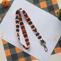 A Halloween-themed fabric lanyard strap with a black metal clasp, featuring a repeating pattern of black bats and ghosts on an orange background, laid flat on a white sheet of paper.
