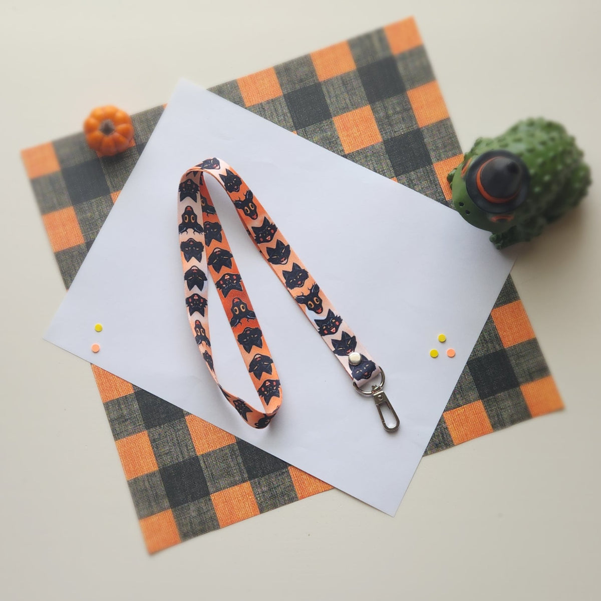 A Halloween-themed fabric lanyard with a black cat pattern on an orange background, laid out on a white sheet of paper over an orange and black plaid mat.