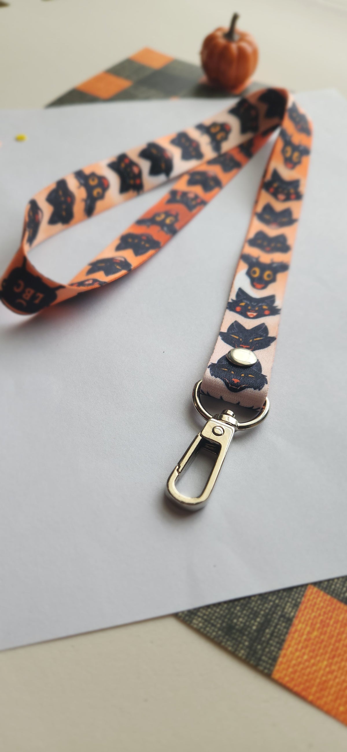 A Halloween-themed fabric lanyard strap featuring a repeating pattern of black cat silhouettes on an orange background, with a silver metal lobster clasp. The strap is laid out on a white sheet of paper with a small decorative pumpkin prop in the background.