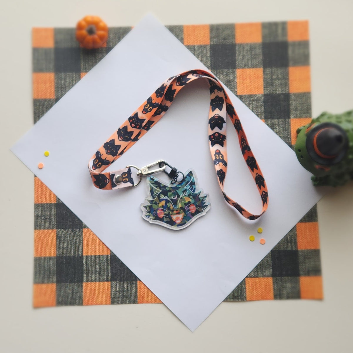 A Halloween-themed keychain or lanyard strap with a colorful cat-shaped charm, displayed on a white card over an orange and black buffalo plaid fabric background with small decorative pumpkins.