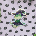 A die-cut sticker featuring a cute green frog dressed as a witch with a purple hat and skirt, placed on a light lavender background patterned with black cat faces and accented by green leaves.
