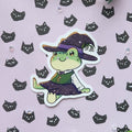 A die-cut sticker featuring a cute green cartoon frog dressed as a witch, placed on a light lavender background patterned with small black cat faces and accented with green leaves.