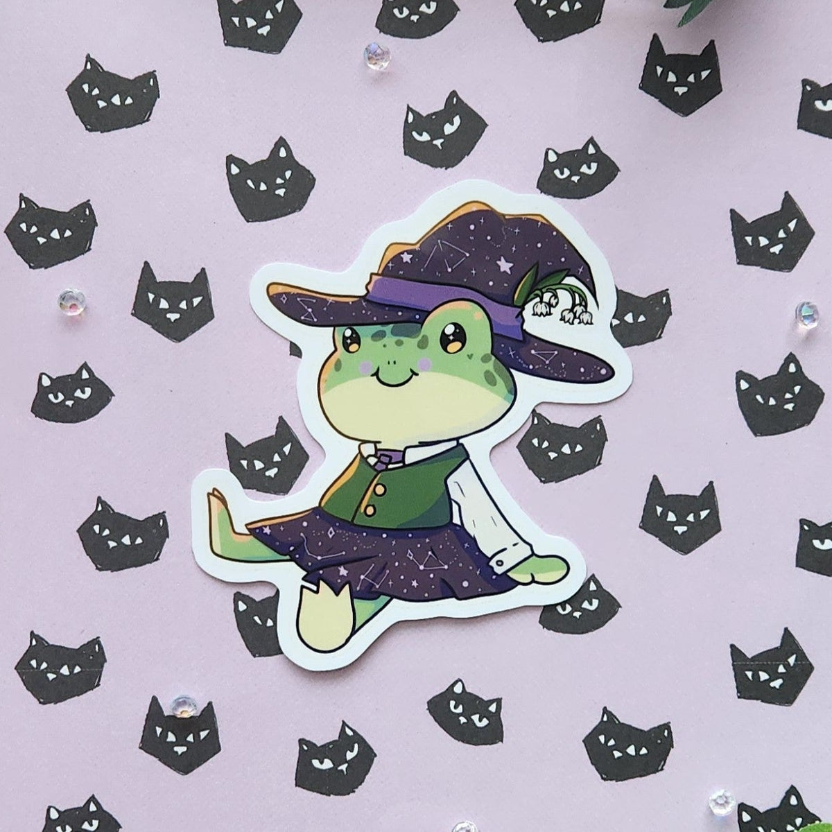 A die-cut sticker featuring a cute green cartoon frog dressed as a witch, placed on a light lavender background patterned with small black cat faces and accented with green leaves.
