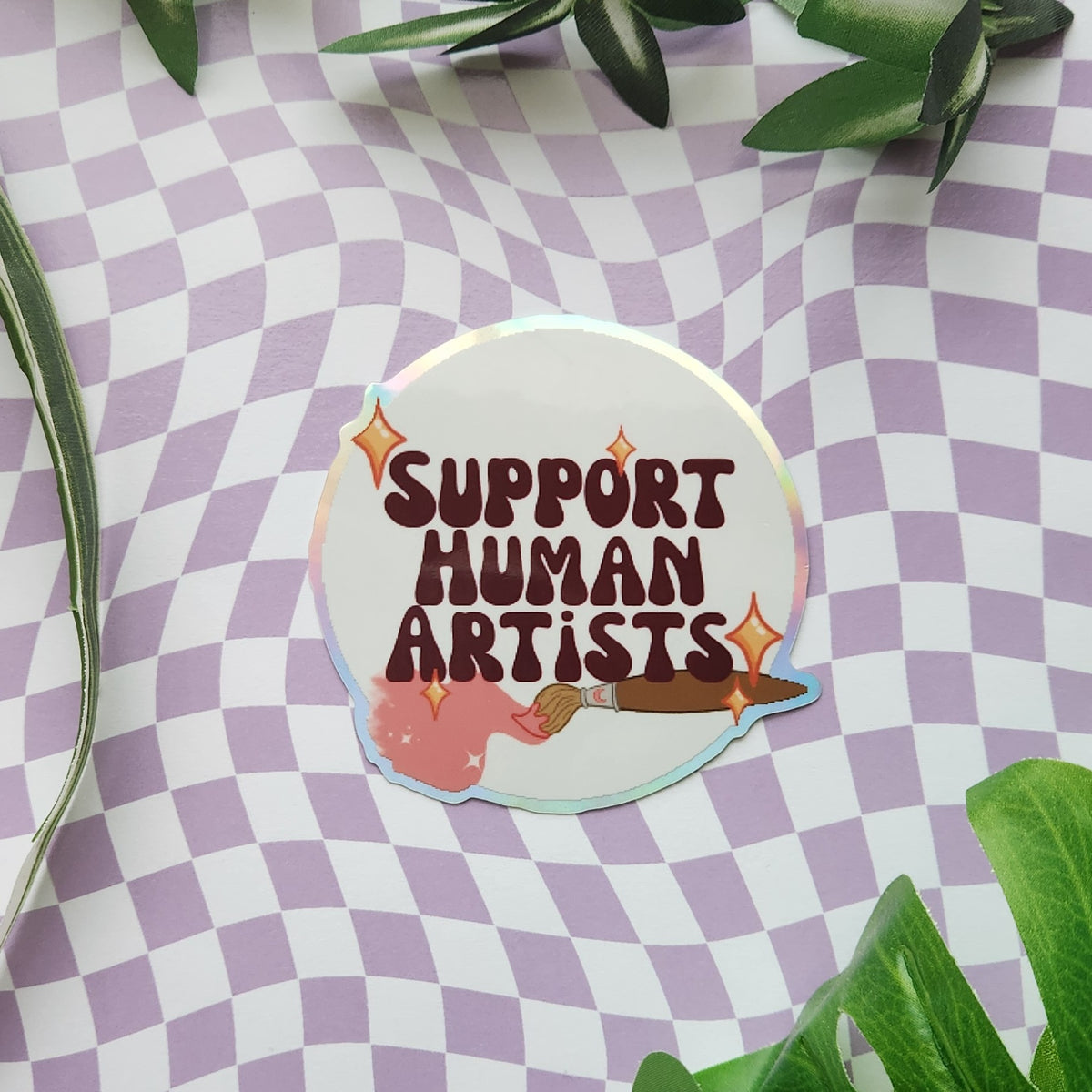 A die-cut sticker with the text 'SUPPORT HUMAN ARTISTS' in dark brown lettering, featuring a paintbrush and paint splatter illustration, placed on a purple and white checkered background with green leaves.