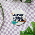 A round enamel pin with the text 'SUPPORT HUMAN ARTISTS' and a paintbrush illustration, displayed on a purple and white checkered background with green leaves.