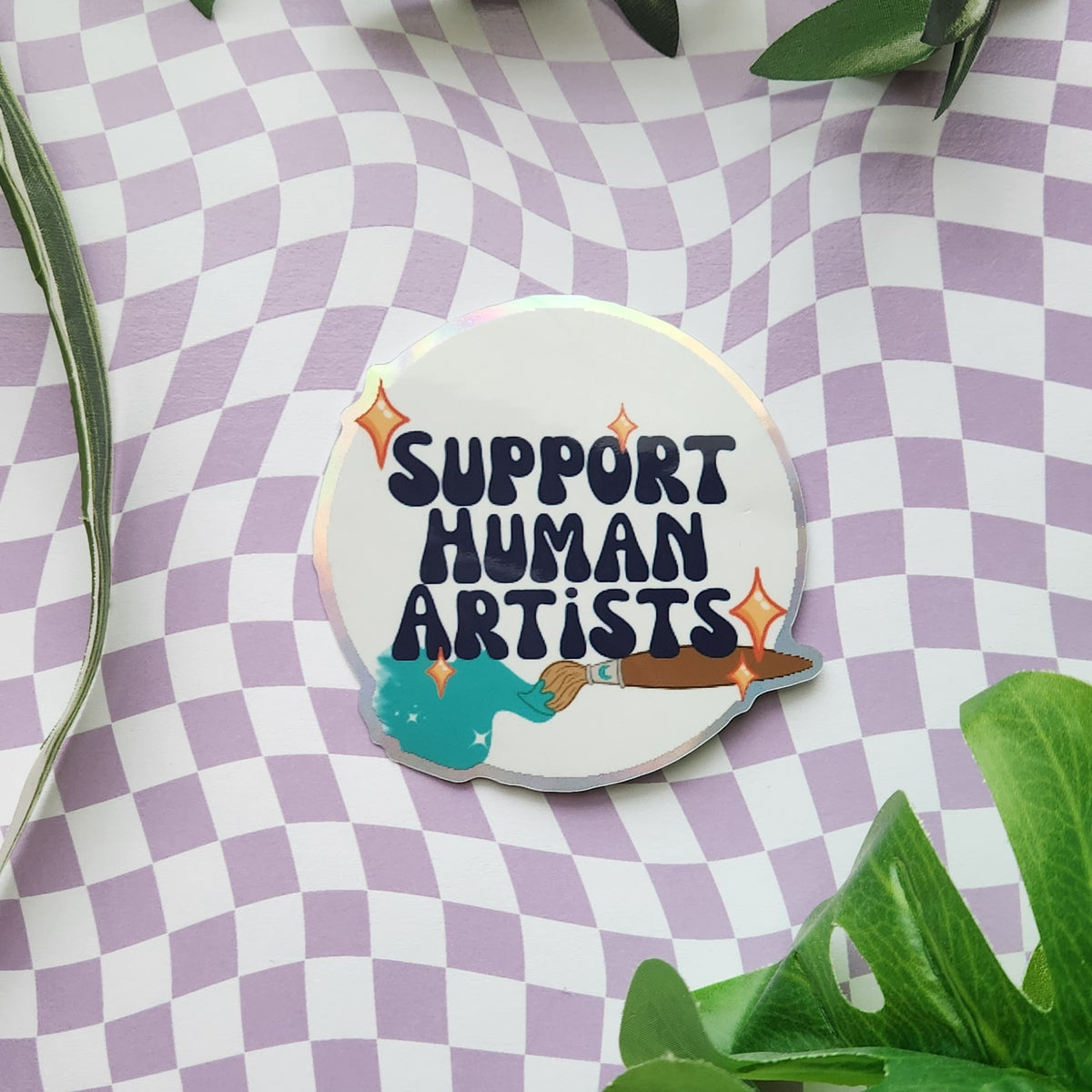 A round enamel pin with the text 'SUPPORT HUMAN ARTISTS' and a paintbrush illustration, displayed on a purple and white checkered background with green leaves.