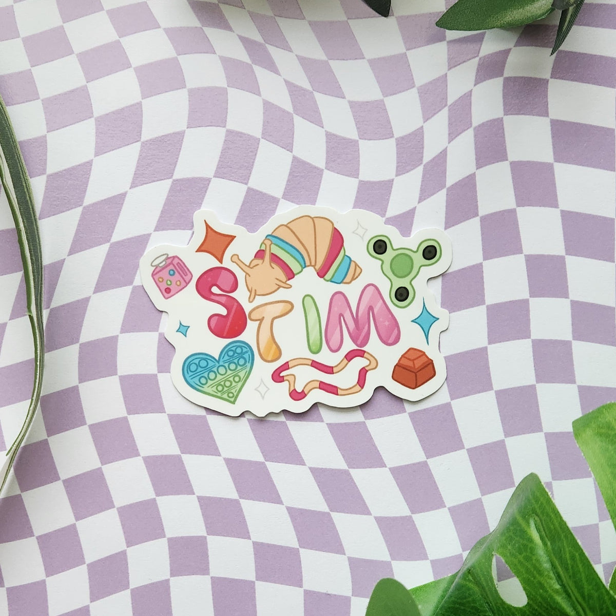 A die-cut sticker featuring the word 'STIM' in colorful retro-style letters with playful doodles, placed on a lavender and white checkered surface with green leaves at the corners.