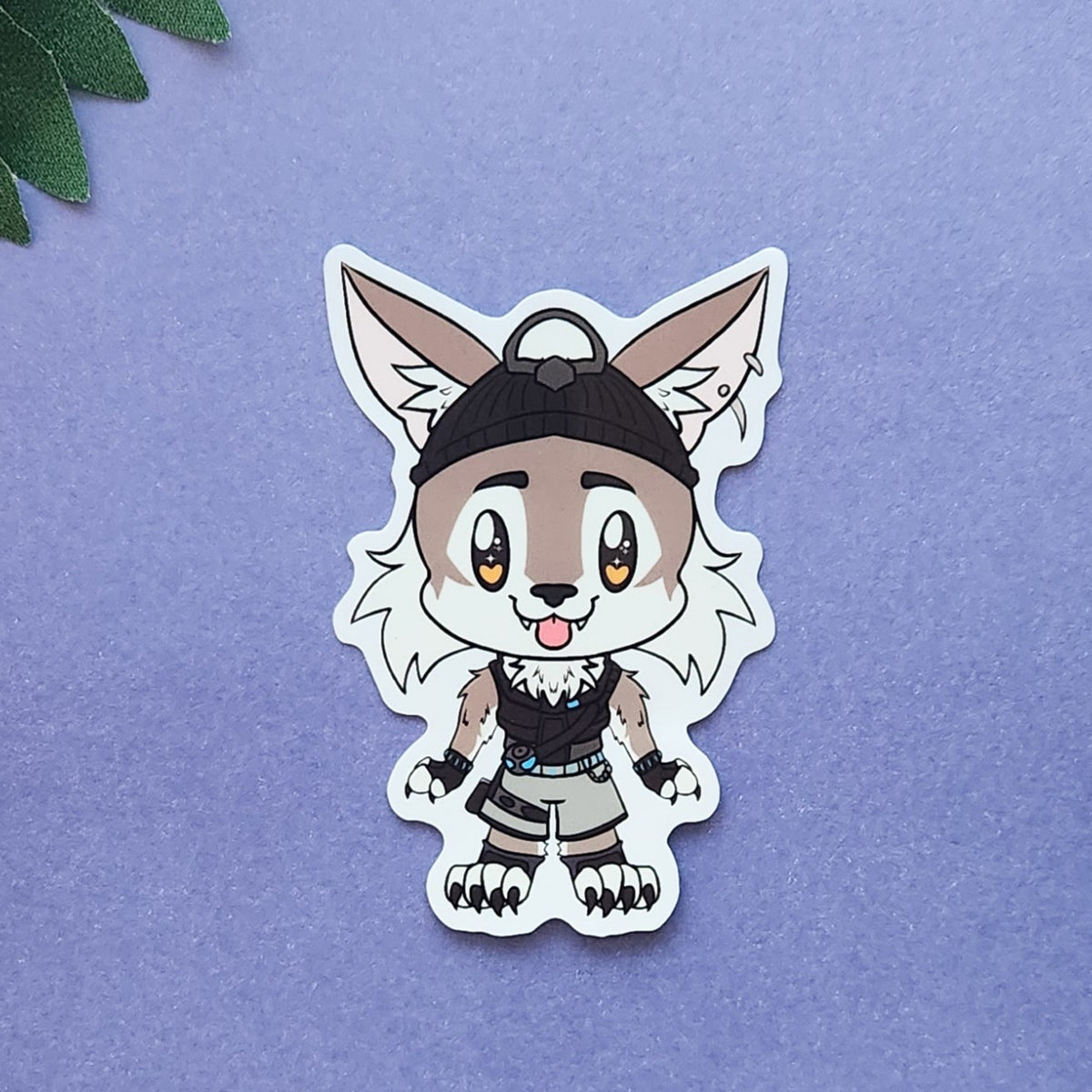 A die-cut vinyl sticker featuring a chibi-style cartoon wolf character wearing a black beanie and dark outfit, placed on a textured lavender background with a small green leaf in the top-left corner.