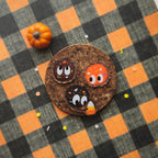 Three Halloween-themed decorative pins shaped like chocolate-covered cookies with cute cartoon faces, arranged on a black and orange plaid fabric backdrop next to a small orange pumpkin.