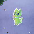 A die-cut sticker featuring a cute green cartoon cat holding an ice cream cone, placed on a textured lavender-blue background with small decorative star and leaf elements.