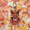 A cartoon-style acrylic keychain charm featuring an angry brown fox with black and orange stripes on its ears and a silver metal clasp, displayed against a watercolor pumpkin patterned background.