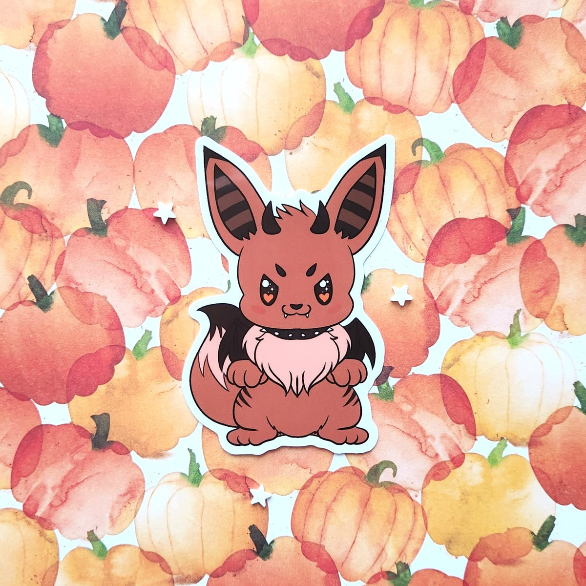 A die-cut sticker featuring a cute cartoon bat fox character, shown against a watercolor pattern of orange and yellow pumpkins.