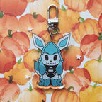 A blue and white skeleton cartoon character keychain charm with a silver lobster clasp, displayed on a watercolor pumpkin patterned background.