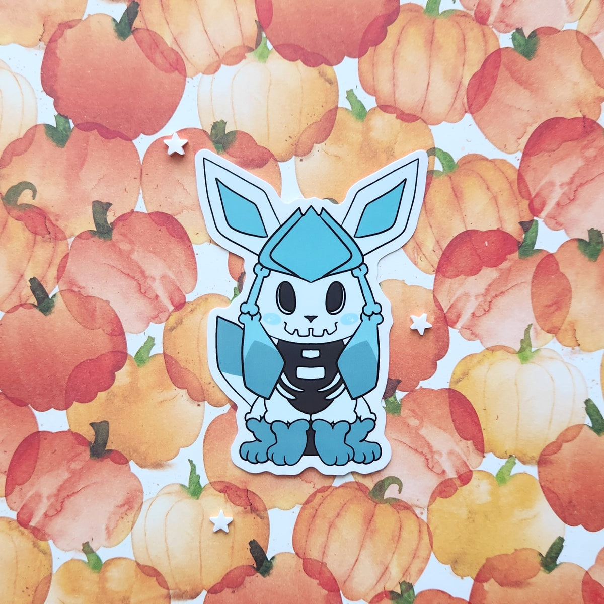 A die-cut sticker featuring a stylized blue and black cartoon fox character, placed on top of a watercolor pumpkin patterned background.