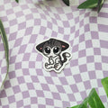 A die-cut sticker featuring a cute cartoon cat with large eyes, sitting on a lavender and white checkered background with green leaves in the corners.