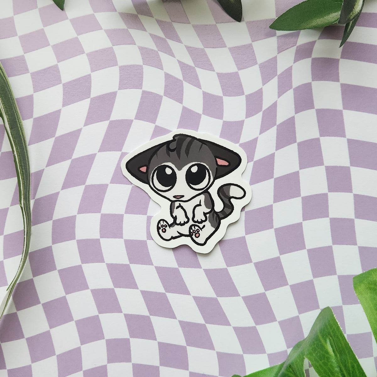 A die-cut sticker featuring a cute cartoon cat with large eyes, sitting on a lavender and white checkered background with green leaves in the corners.