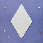 A translucent white diamond-shaped decorative plaque with embossed floral patterns, displayed on a textured purple background with small scattered star and moon embellishments.