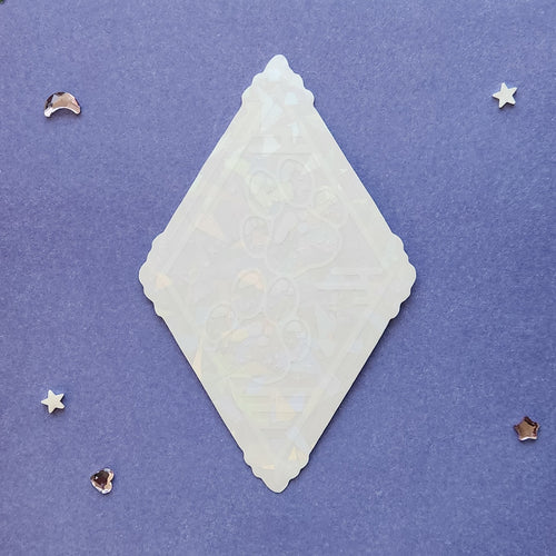 A translucent white diamond-shaped decorative plaque with embossed floral patterns, displayed on a textured purple background with small scattered star and moon embellishments.