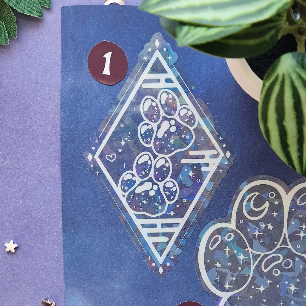 A close-up of a decorative sticker or decal sheet featuring a large diamond-shaped paw print design with a galaxy pattern and white border, placed on a purple background with plant leaves and small decorative stars.