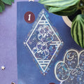 A close-up of a decorative sticker or decal sheet featuring a large diamond-shaped paw print design with a galaxy pattern and white border, placed on a purple background with plant leaves and small decorative stars.
