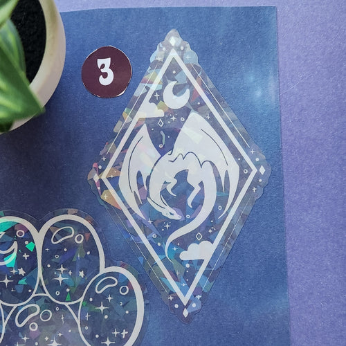 A holographic diamond-shaped sticker featuring a white dragon design, displayed on a blue background alongside other stickers. A small plant pot is visible in the top-left corner.