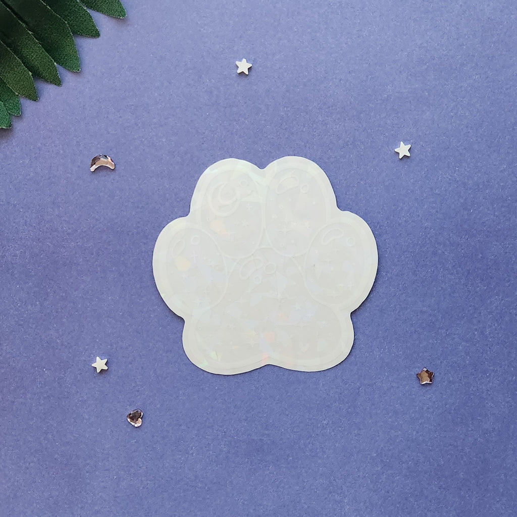 A translucent, paw-shaped acrylic nail art sticker or decal with a subtle iridescent sheen, displayed on a textured purple background with decorative star and leaf props.