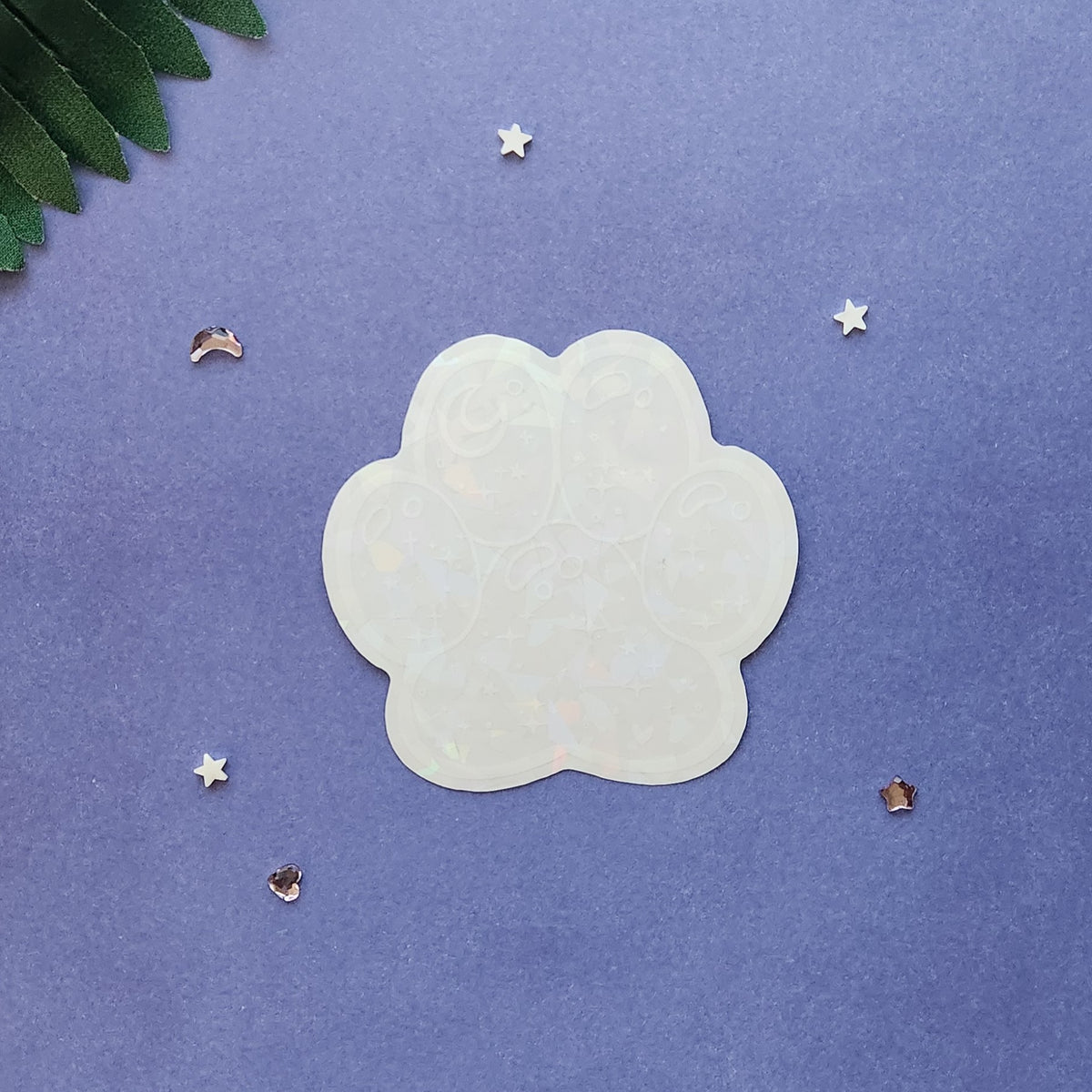 A translucent, paw-shaped acrylic nail art sticker or decal with a subtle iridescent sheen, displayed on a textured purple background with decorative star and leaf props.
