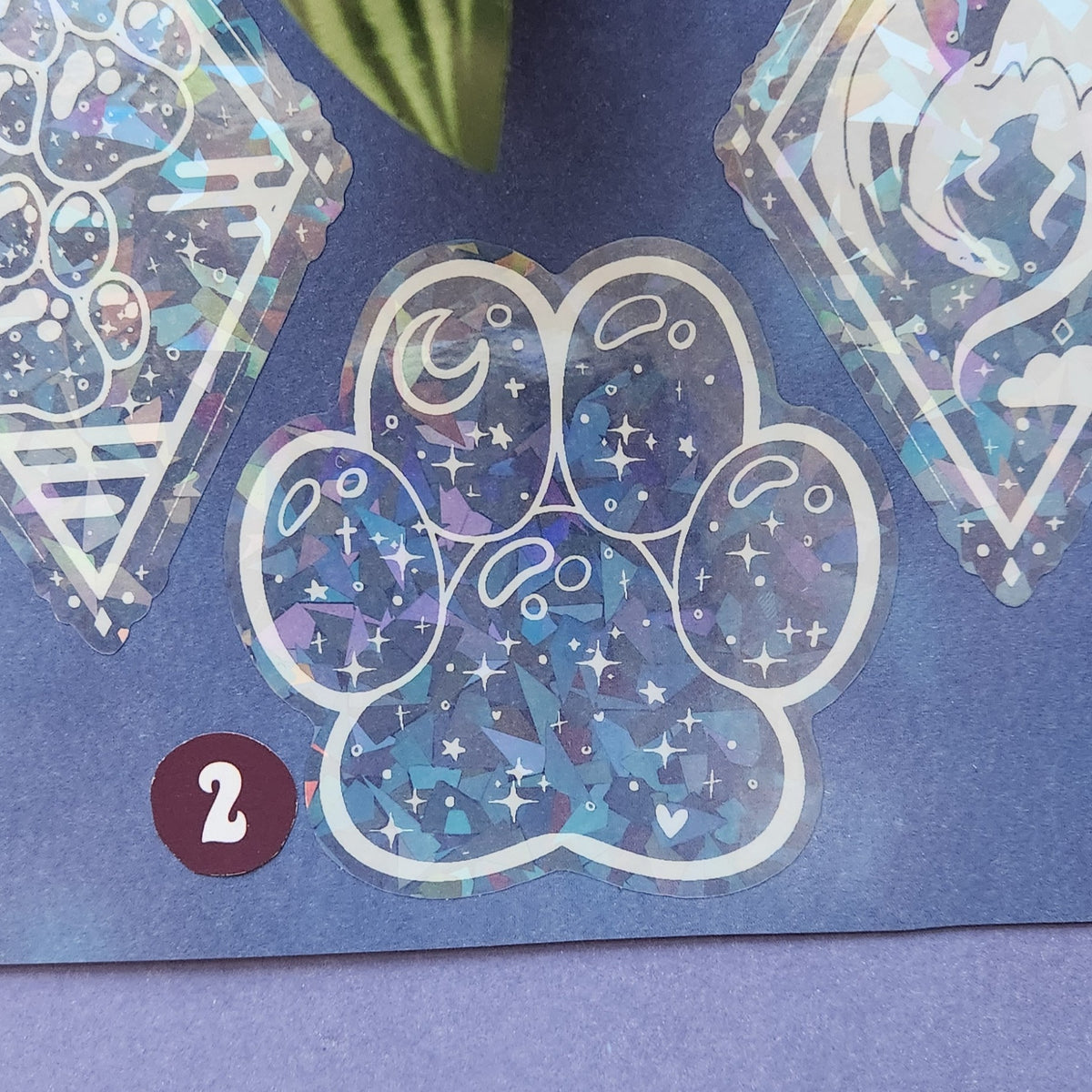 A close-up of a paw-shaped sticker or decal with a white outline and a blue-purple holographic glitter pattern featuring stars, moons, and small hearts. The sticker is placed on a dark blue surface, with partial other decorative stickers visible around it and a small maroon circle labeled '2'.