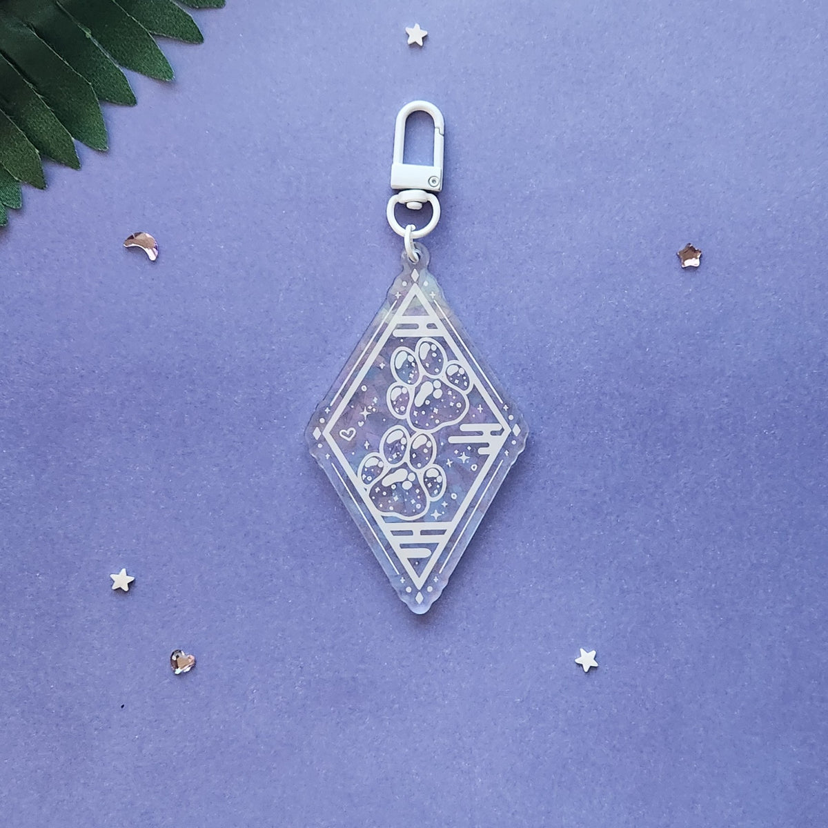 A clear acrylic keychain charm with a silver clasp, featuring a paw print design with glitter and small star details, displayed on a textured purple background with decorative star confetti and a green leaf.