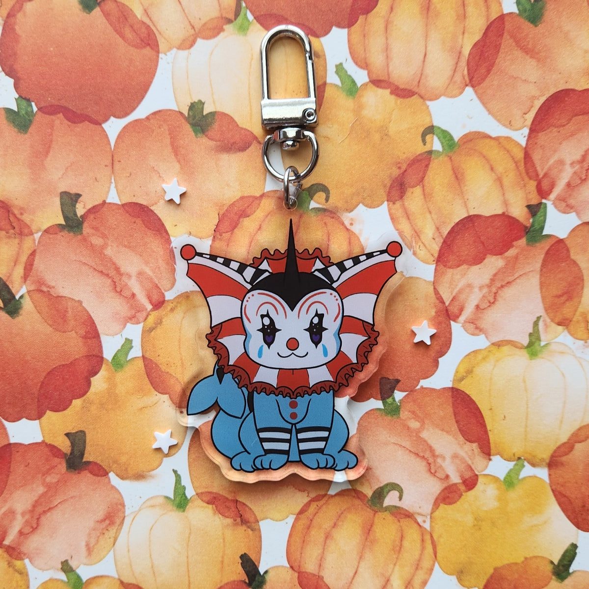 A cartoon jester-themed keychain charm with a silver clasp, displayed on a watercolor pumpkin patterned background.