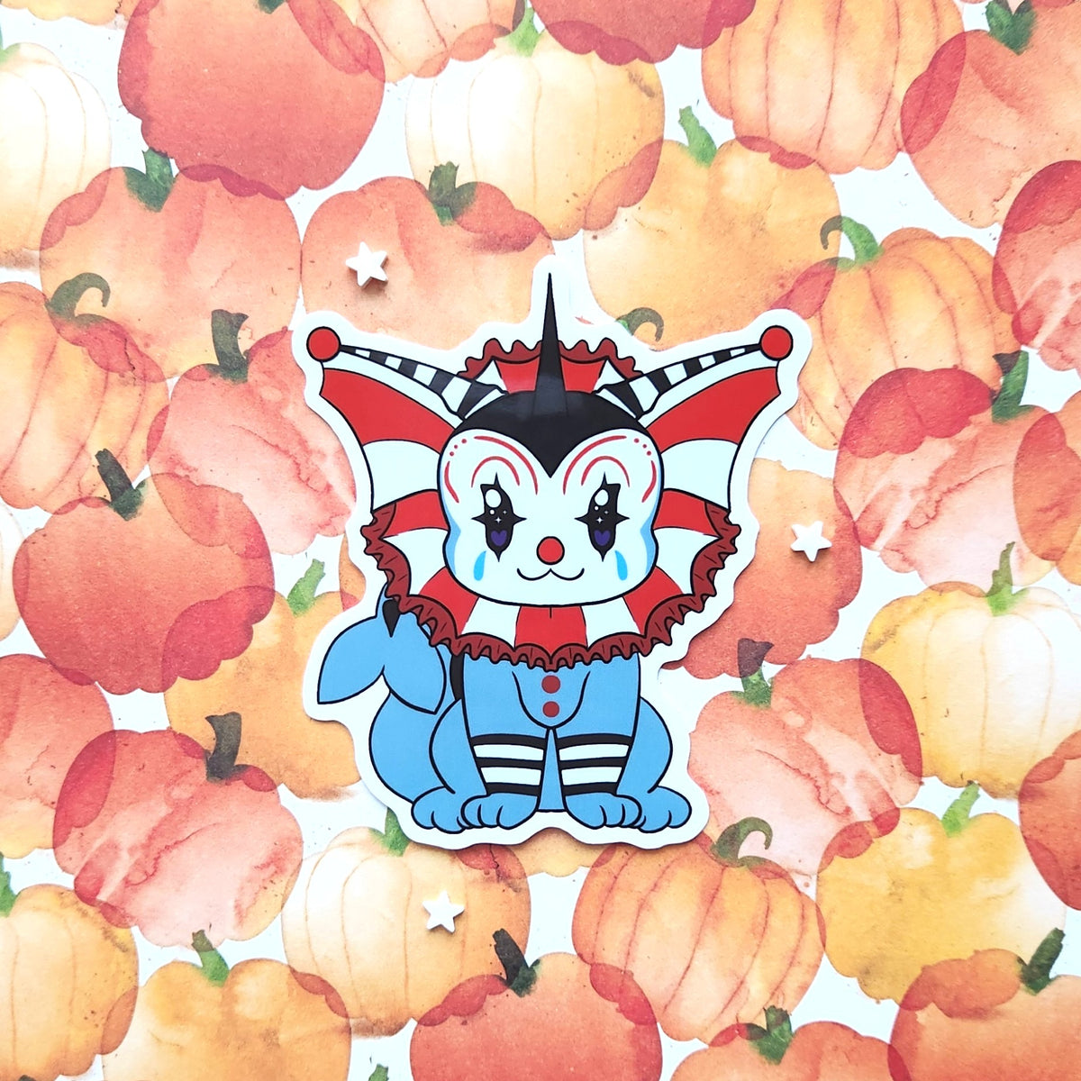 A die-cut sticker featuring a cute cartoon clown character with a red, white, and black jester hat, blue outfit, and red nose, placed on top of a watercolor pumpkin patterned background.