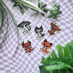 Five cute cartoon animal stickers with white die-cut borders are arranged on a lavender-and-white checkered sheet with green plant leaves around them. The animals include a black-and-white cat, a husky puppy, a brown deer, a red dragon, and an orange fox.
