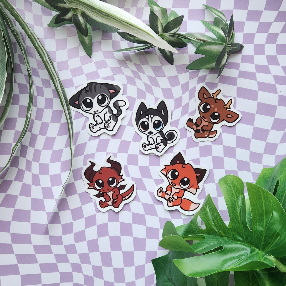 Five cute cartoon animal stickers with white die-cut borders are arranged on a lavender-and-white checkered sheet with green plant leaves around them. The animals include a black-and-white cat, a husky puppy, a brown deer, a red dragon, and an orange fox.