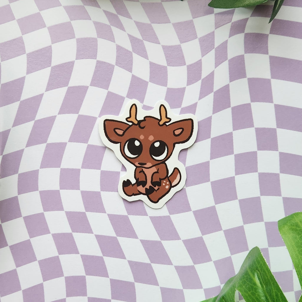 A die-cut sticker featuring a cute cartoon brown deer with large eyes and small antlers, placed on a lavender and white wavy checkered background with green leaves in the corners.