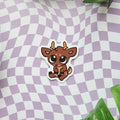 A die-cut sticker featuring a cute cartoon brown deer with large eyes and small antlers, placed on a lavender and white wavy checkered background with green leaves in the corners.