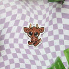 A die-cut sticker featuring a cute cartoon brown deer with large eyes and small antlers, placed on a lavender and white wavy checkered background with green leaves in the corners.