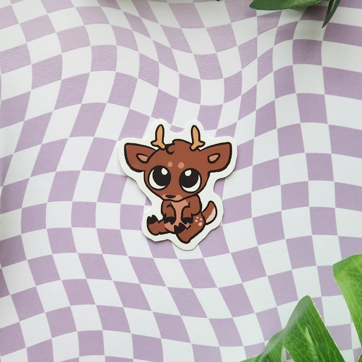 A die-cut sticker featuring a cute cartoon brown deer with large eyes and small antlers, placed on a lavender and white wavy checkered background with green leaves in the corners.