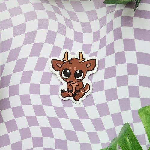 A die-cut sticker featuring a cute cartoon brown deer with large eyes and small antlers, placed on a lavender and white wavy checkered background with green leaves in the corners.