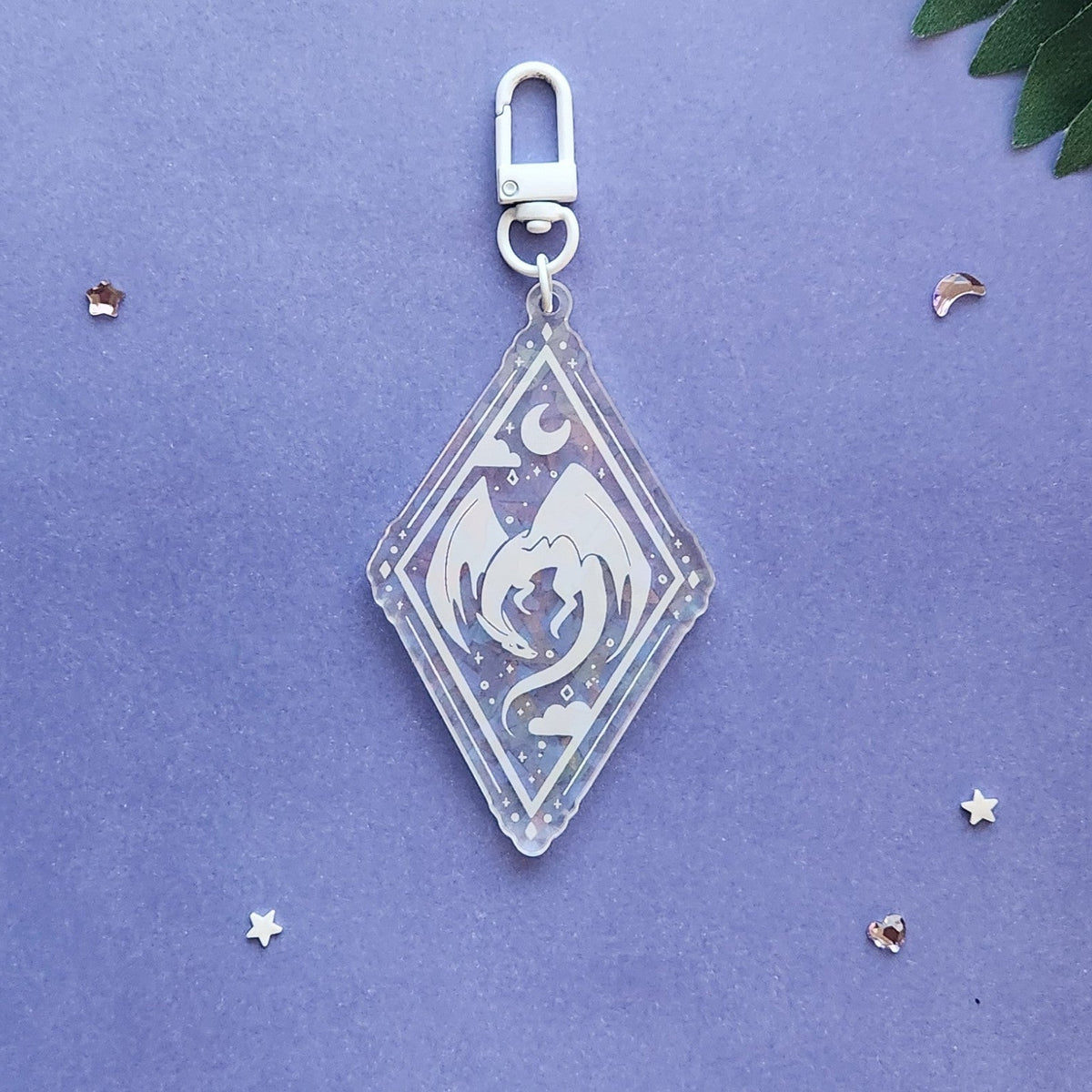 A clear acrylic keychain charm with a silver clasp, featuring a white dragon design, displayed on a textured purple background with small decorative star and leaf props.