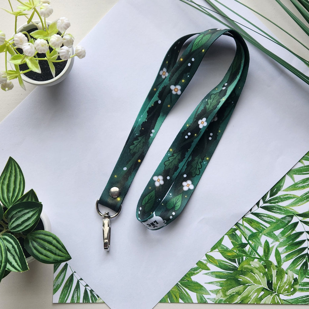 A dark green printed lanyard with a forest pattern of leaves and small white flowers, laid flat on a white sheet of paper with decorative plants around it.
