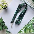 A dark green printed lanyard with a forest pattern of leaves and small white flowers, laid flat on a white sheet of paper with decorative plants around it.