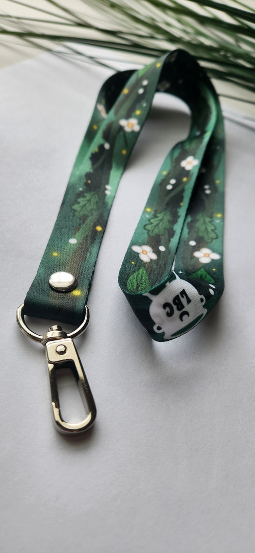 A dark green printed lanyard with a woodland pattern of leaves and small white flowers, featuring a silver metal carabiner clasp and a white tag with black text.