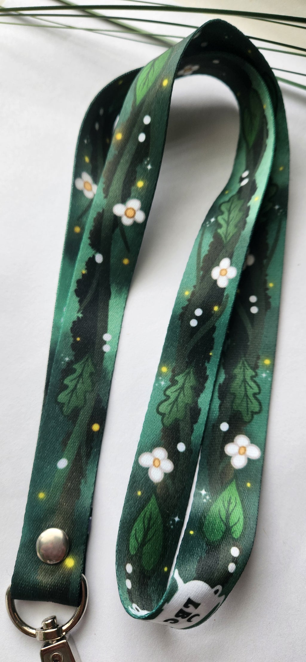 A dark green fabric lanyard with a printed pattern of white flowers, green leaves, and small yellow dots, featuring a silver metal snap button and keyring clasp.