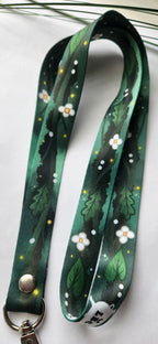 A dark green fabric lanyard with a printed pattern of white flowers, green leaves, and small yellow dots, featuring a silver metal snap button and keyring clasp.