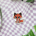 A die-cut sticker featuring a cute cartoon fox in orange, brown, and black, placed on a lavender and white checkered surface with green leaves in the corners.