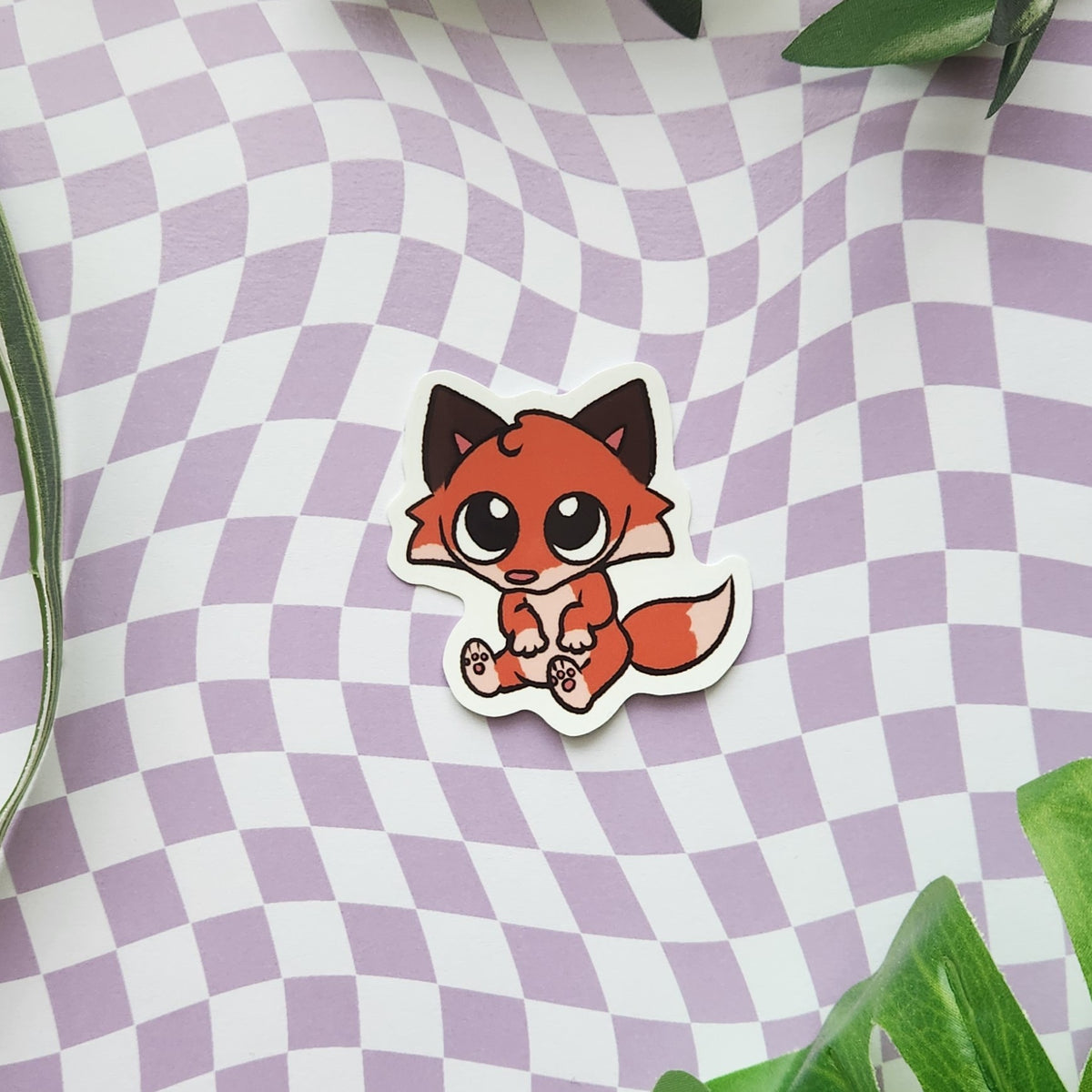 A die-cut sticker featuring a cute cartoon fox in orange, brown, and black, placed on a lavender and white checkered surface with green leaves in the corners.