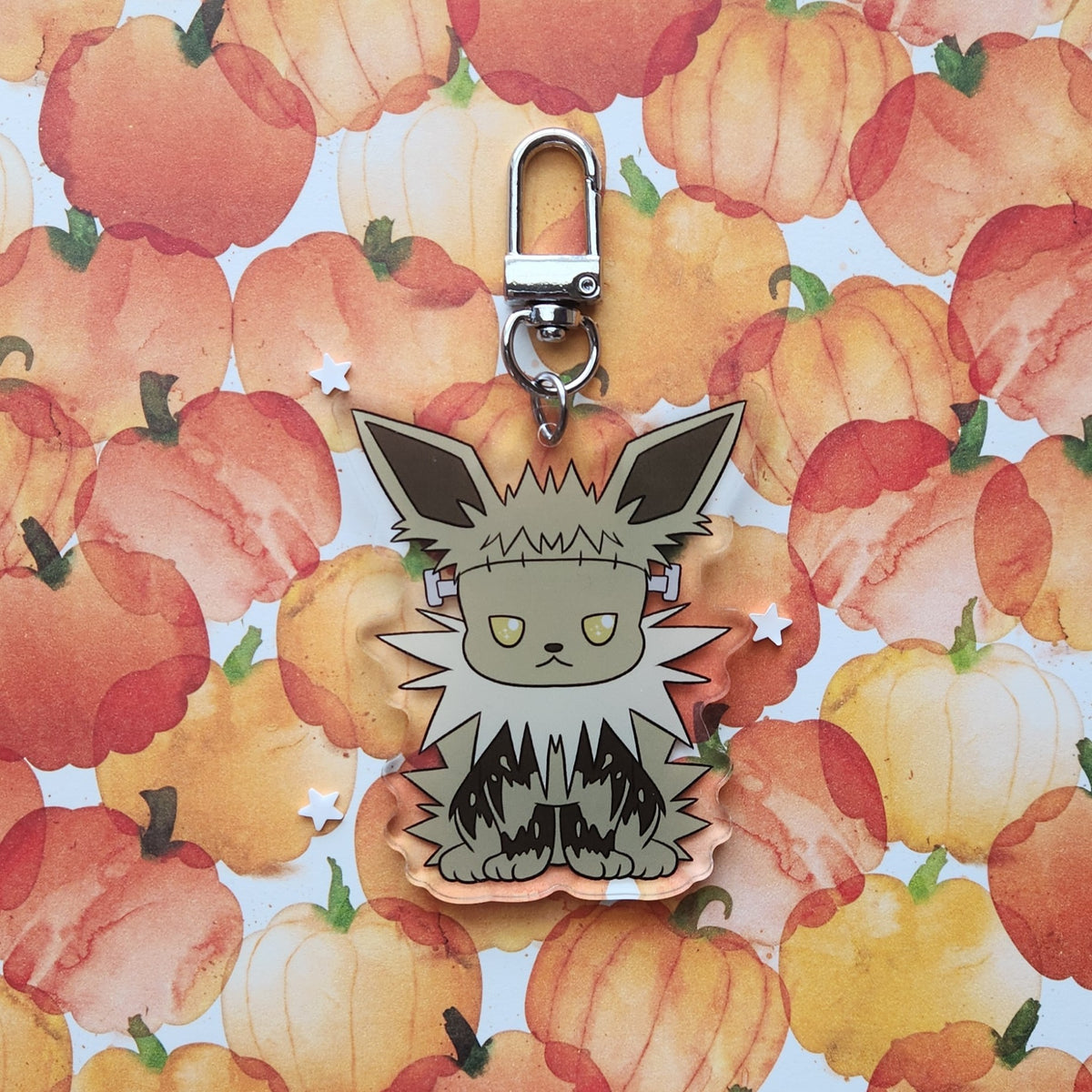 A character keychain featuring a stylized gray and black furry creature with yellow eyes, displayed against a watercolor pumpkin patterned background.