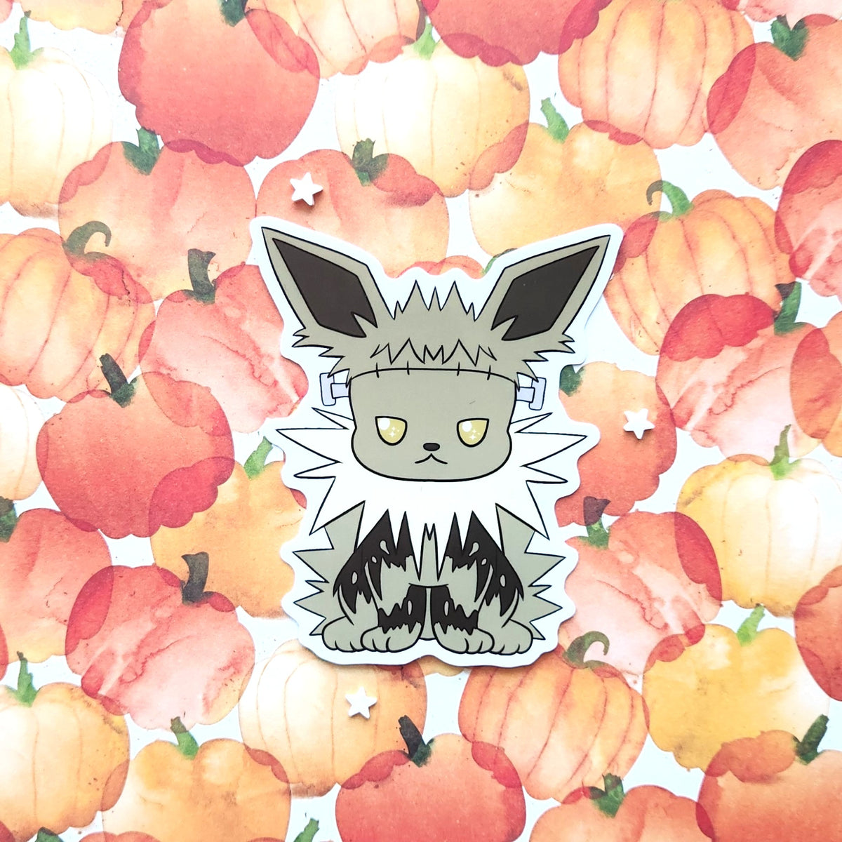 A die-cut sticker featuring a cute cartoon character with large pointed ears, yellow eyes, and black-and-white striped legs, placed on a watercolor pumpkin patterned background.