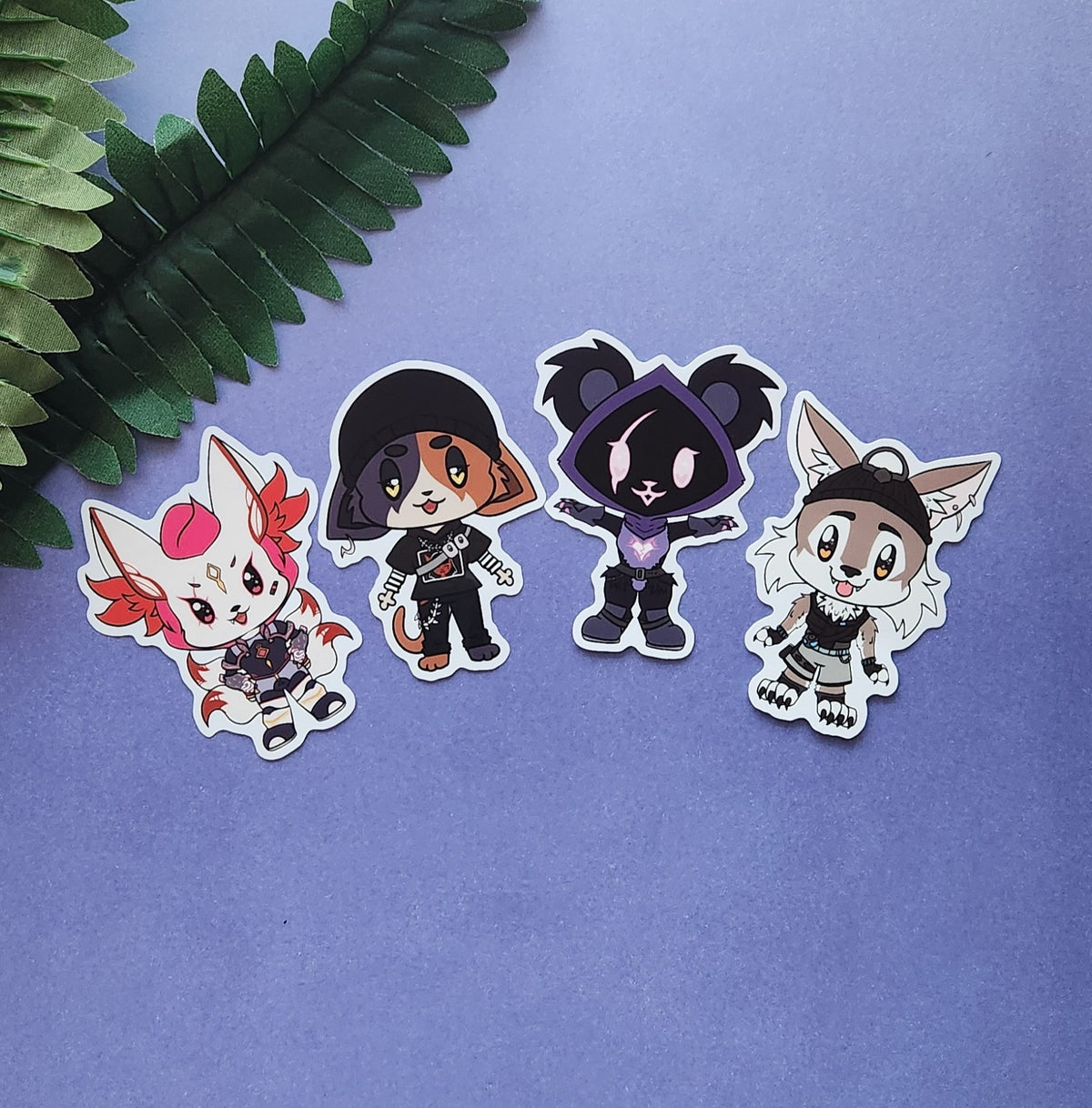 Four die-cut vinyl stickers featuring cute chibi anime-style animal characters, arranged on a textured lavender-blue background with a green fern leaf in the top-left corner.