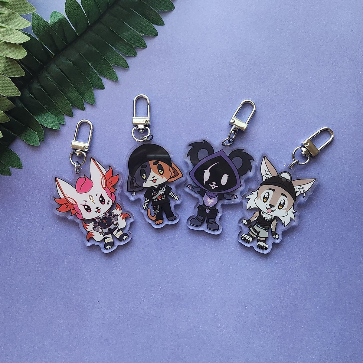 Four chibi anime keychains with silver lobster clasps, arranged on a lavender background with a green fern leaf in the top left corner.